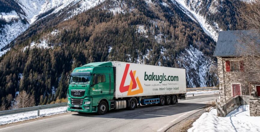 A bakugls.com lorry driving on a road near a tall snowy mountain with trees on it. Next to the road there is a house made of bricks with red window shutters and a red door